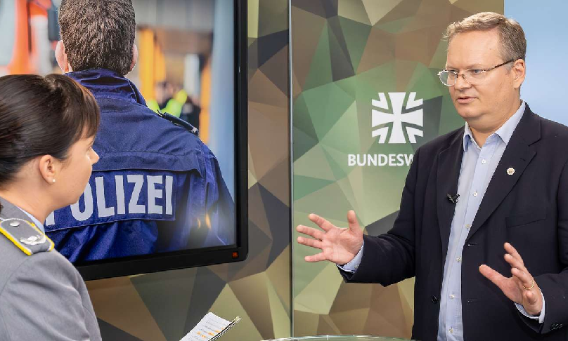 Foto: Bundeswehr/Steve Eibe BAKS-Vizepräsident Markus Woelke spricht mit einer Soldatin im Dienstgrad Major der Bundeswehr vor einem Bildschirm, der einen Polizisten und einen Rettungswagen zeigt.