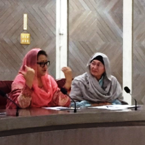 Zwei Frauen in traditioneller islamischer Kleidung sitzen an einem Tisch mit Mikrofonen; eine spricht, die andere blickt zu ihrer Nachbarin.