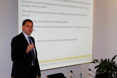 Ein Mann im dunklen Anzug steht gestikulierend vor einer Leinwand und hält einen Vortrag.