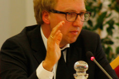Ein Mann sitzt an einem Tisch und gestikuliert mit der rechten Hand.