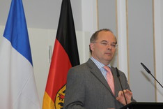 Ein Herr im grauen Anzug steht an einem Rednerpult. Im Hintergrund sind die französische und die deutsche Flagge zu sehen.