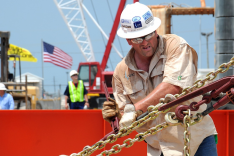 Ein Mann in Arbeitskleidung und Schutzhelm hantiert mit Werkzeug an einer gespannten Kette; im Hintergrund weht eine US-amerikanische Flagge, und weiteres Arbeitsgerät ist zu sehen.