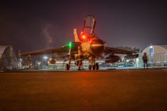 Ein Kampfflugzeug vom Typ Tornado steht bei Nacht auf einem beleuchteten Vorfeld zwischen zwei Hangars.