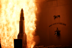 Eine Rakete verlässt vor einem Feuerschweif einen Startkanister eines Kriegsschiffs; daneben ist eine Schiffsglocke mit dem Motto des Schiffs erkennbar.