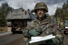 Ein Bundeswehrsoldat in Kampfausrüstung steht hinter dem Heck einer Panzerhaubitze 2000; in den Händen hält er eine Karte und ein Funkgerät.
