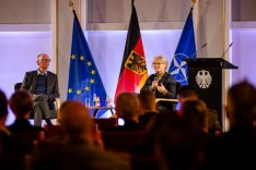 BAKS-Präsident Brose und Verteidigungsministerin Lambrecht sitzen vor der EU-, der Bundes- und der NATO-Flagge und sprechen zum Publikum im Vordergrund.