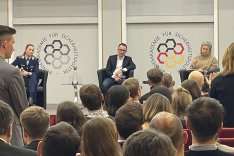 Eine Soldatin und zwei Zivilpersonen sitzen auf einem Podium und blicken zu einem Publikum, aus dem ein Mann und eine Frau stehend mit ihnen interagieren.