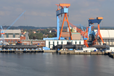 Auf einem Werftgelände am Wasser liegen mehrere Unterseeboote im Bau an Land; darüber ragen große Werftkräne auf.