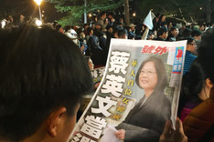 Ein Mann blickt am Tag der taiwanesischen Wahl auf eine Zeitung, die Tsai Ing-wen abbildet, die Spitzenkandidatin der DPP bei den Präsidentschaftswahlen. Im Hintergrund stehen Menschen mit Fahnen. 