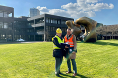 Foto: BAKS/Beu Vor der Fassade des Bundeskanzleramts in Bonn stehen zwei Männer mit Warnwestern, einer mit Bauhelm auf einer Rasenfläche; rechts ist die Skulptur Large Two Forms des Künstlers Henry Moore erkennbar..