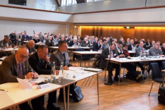 Männer, darunter einige in militärischer Uniform, sitzen in einem großen Saal an langen Tischen.