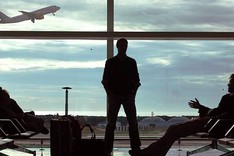 Zwei Personen, eine stehend, die andere sitzend, im Wartebereich eine Flughafens. Durch die Großen Fenster sieht man ein startendes Passagierflugzeug.