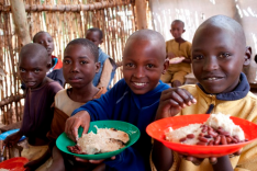 Eine Gruppe afrikanischer Kinder sitzt mit gefüllten Tellern in den Händen in einem Gebäude aus weidengeflechtartigen Holzwänden und blickt zum Betrachter.