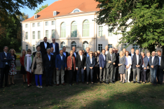 Eine Gruppe geschäftlich gekleideter Menschen steht auf dem Rasen zwischen Bäumen vor einem Schloss.