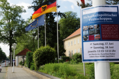 Vor dem Haus Berlin der BAKS wehen die Flaggen der EU, Deutschlands und Berlins; an einem Laternenmast recht hängt ein Plakat mit der Aufschrift &quot;Sicherheitspolitischer Frühschoppen&quot;.