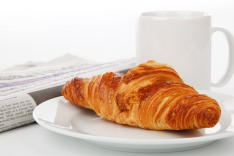 Foto eines Croissants vor einer Kaffeetasse. Im Hintergrund ist unscharf eine Zeitung zu sehen.