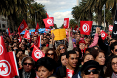Auf einer Straße gesäumt von Palmen steht eine große Menschenmenge, die zahlreiche Flaggen Tunesiens und Plakate in arabischer Sprache mit sich führt.