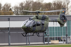 Ein Hubschrauber vom Typ Airbus H145M in Tarnlackierung der Bundeswehr fliegt niedrig vor einem Hangargebäude.