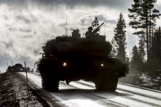 Schemenhaft sind zwei Panzerfahrzeuge auf einer Straße zu erkennen; rechts ragen Nadelbäume auf.