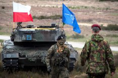 Ein Soldat aus Polen und ein Soldat aus den Niederlanden stehen sich vor einem Panzer, auf dem die Flaggen Polens und der NATO gehisst sind, gegenüber.