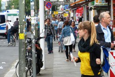 Zahlreiche Menschen stehen und gehen entlang einer Einkaufsstraße in Berlin-Neukölln.