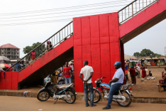 Zahlreiche afrikanische Frauen und Männder stehen und sitzen bei einem nach oben rechts aus dem Bild ragenden rot gestrichenen Treppenaufgang aus Beton. Vorn stehen zwei Krafträder; im Hintergrund ragt links ein Neubau, rechts improvisierte Stände auf.