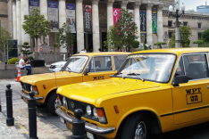 Im Vordergrund stehen zwei gelbe Taxis osteuropäischer Bauart; im Hintergrund ragt eine Säulenkollonade mit Plakaten zwischen den Säulen auf.