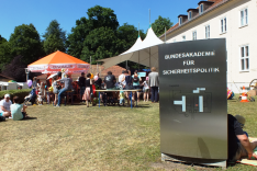 Auf der Wiese vor dem Haus Bonn der BAKS stehen Zelte und eine Menschenmenge, letztere zum Teil sitzend auf Bänken und dem Boden; rechts ragt das Haus selbst auf; im Vordergrund rechts steht ein großes Metallschild mti dem Namen der BAKS.