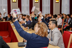 In einem Konferenzsaal sitzen geschäftlich gekleidete junge Menschen an Konferenztischen und halten Tischschilder mit den Namen und Flaggen von NATO-Mitgliedsstaaten hoch.