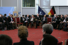 A large group of both uniformed and business-dressed people are sitting in a large circle of chairs within the Federal Academy&#039;s Historic Hall.