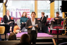 Jana Puglierin spricht mit einem Blatt Papier in der Hand vor einem rundlich angeordneten Diskussionspodium mit vier sitzenden Beteiligten stehen zur betrachtenden Person.