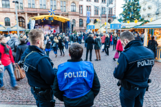 Drei Polizisten stehen bei Tag auf einem Weihnachtsmarkt während zahlreiche Passanten vorbeigehen.