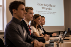 Vier junge Menschen sitzen an einem Tisch; im Hintergrund wird eine Präsentation mit dem Titel "Cyberangriffe auf Wikipedia"  auf eine Leinwand projiziert.