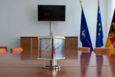 Auf einem Konferenztisch steht eine Uhr; im Hintergrund stehen drei Flaggen und ein Bildschirm hängt an einer weißen Wand.