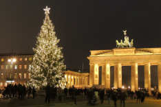 Das Foto das das Brandenburger Tor in Berlin bei Nacht von Osten aus betrachtet; es ist ebenso wie ein Gebäude daneben und ein großer Weihnachtsbaum davor hell erleuchtet und von zahlreichen Menschen umgeben.