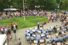 Auf einem ovalen Platz spielt ein uniformiertes Orchester und zahlreiche Menschen haben sich versammelt um zuzuhören.