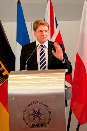 Dr. Ackermann, ein Herr im dunklen Anzug, steht an einem silbernen Rednerpult.