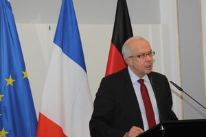 Ein Herr im dunklen Anzug steht an einem Rednerpult. Im Hintergrund sind die französische und die deutsche Flagge zu sehen.
