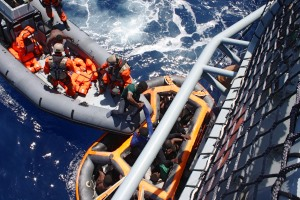 Ein Festrumpfschlauchboot mit Soldaten und eine Rettungsinsel voller Menschen liegen an der Bordwand eines Militärschiffs.