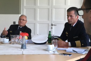 Zwei Männer in Marineoffizieruniformen sitzen an einem Tisch mit Tagungsunterlagen und sprechen zum Betrachter.
