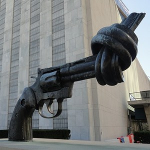 Die metallene Skulptur einer Pistole mit verknotetem Lauf steht vor einem großen, grauen Gebäude.