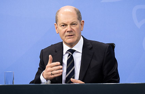 Bundeskanzler Olaf Scholz sitzt an einem Tisch vor einer blauen Pressewand und spricht gestikulierend zur betrachtenden Person.