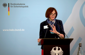 Bettine Cadenbach steht an einem schwarzen Rednerpult mit Bundesadler darauf vor einer blauen Wand mit dem Logo der BAKS und spricht in ein Mikrofon.