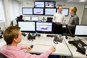 Ein Mann sitzt an einem Computer mit zahlreichen Bildschirm; dahinter stehen zwei weitere Männer, und an der Wand hängen weitere Bildschirme.