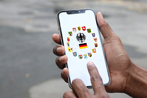 Die Hände einer Person halten ein Smartphone, auf dessen Bildschirm die Wappen der 16 deutschen Bundesländer, ein Bundesadler und die Deutschlandflagge angezeigt werden.