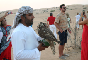 Das Bild zeigt einen arabishen Falkner, der mit westlich gekleideten Menschen in der Wüste steht. 
