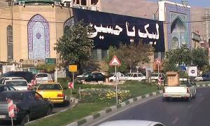 Im Vordergrund zeigt das Bild dichten Autoverkehr, im Mittelgrund ragen Geäbude im islamischen Baustil auf, daran ist ein sehr großes Banner mit Aufschrift in Persisch befestigt; im Hintergrund bildet ein Gebirgskamm den Horizont.