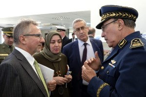 Ein Mann in geschäftlicher Kleidung und ein Mann in Uniform sprechen miteinander; im Hintergrund stehen ein weiterer Mann und eine Frau.