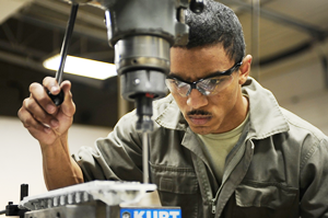 Ein Mann mit Schutzbrille und Arbeitskleidung bedient eine in einen Ständer eingespannte Bohrmaschine und blickt konzentriert auf das zu durchbohrende Werkstück.