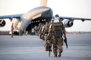 Mehrere Soldaten laufen auf einem Flugfeld mit Gepäck und Waffen in Richtung der geöffneten Heckklappe eines militärischen Transportflugzeugs.